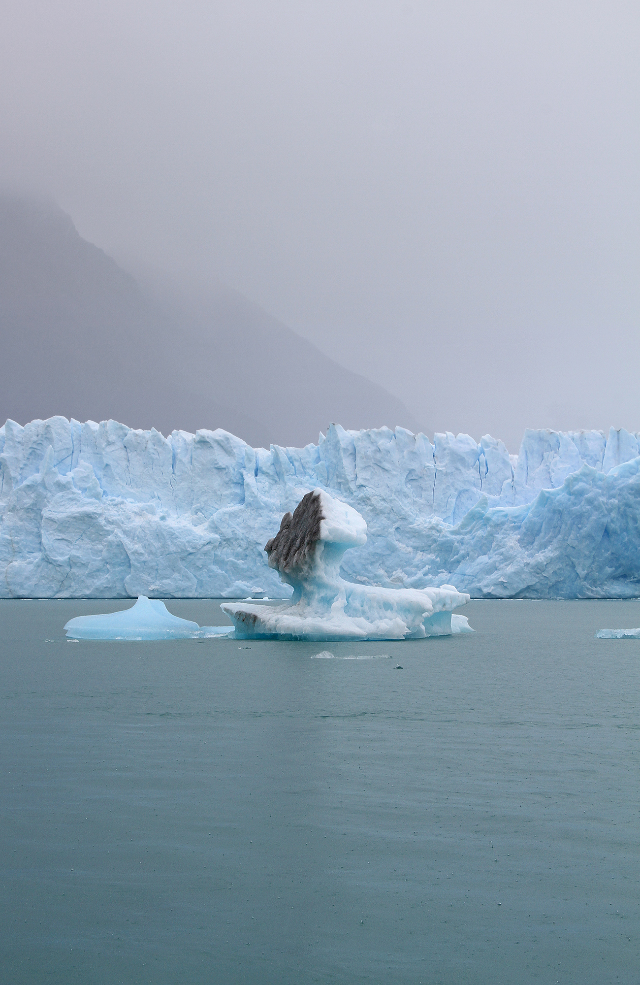 Broken off piece of ice in the middle of a lake, with a massive glacier in the background covered in fog.