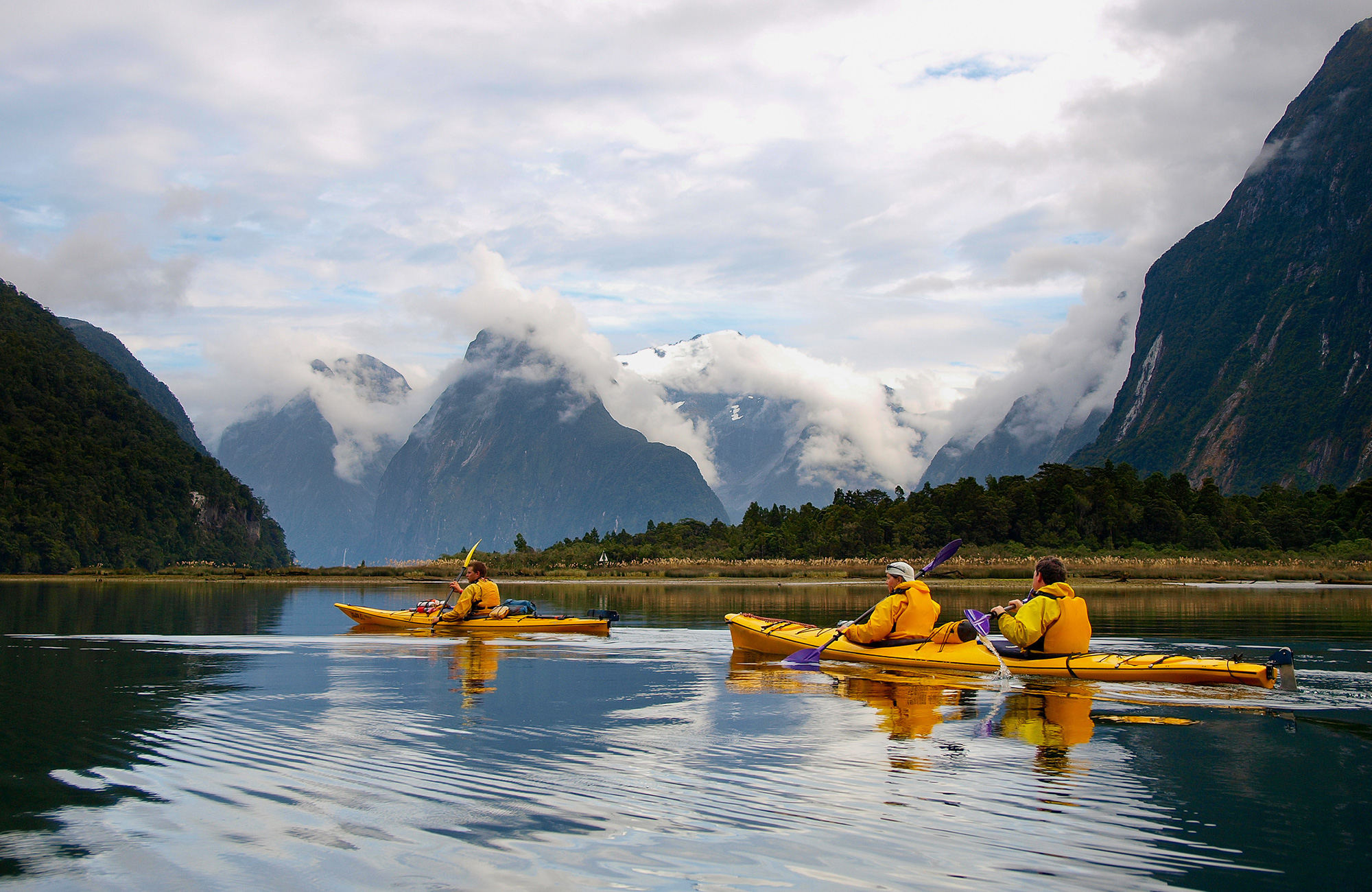 Three people in orange kayaks paddling on a lake in New Zealand with mountains and clouds in the background