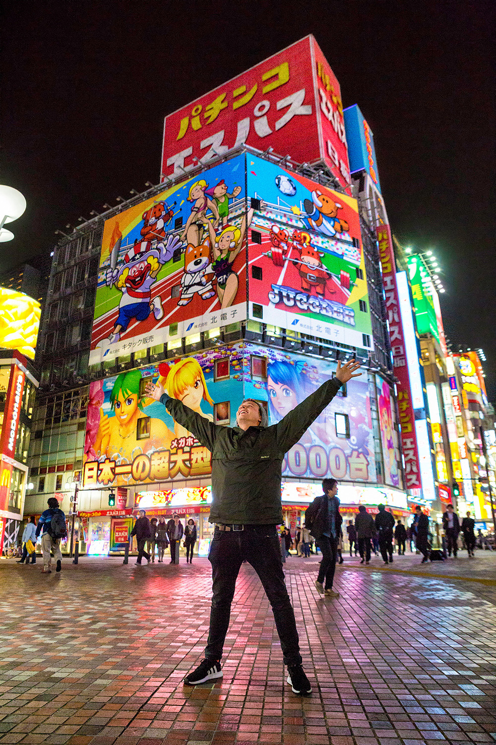 Guy spreading his arms on a square in Tokyo with big billboards with Japanese animation figures on buildings in the background
