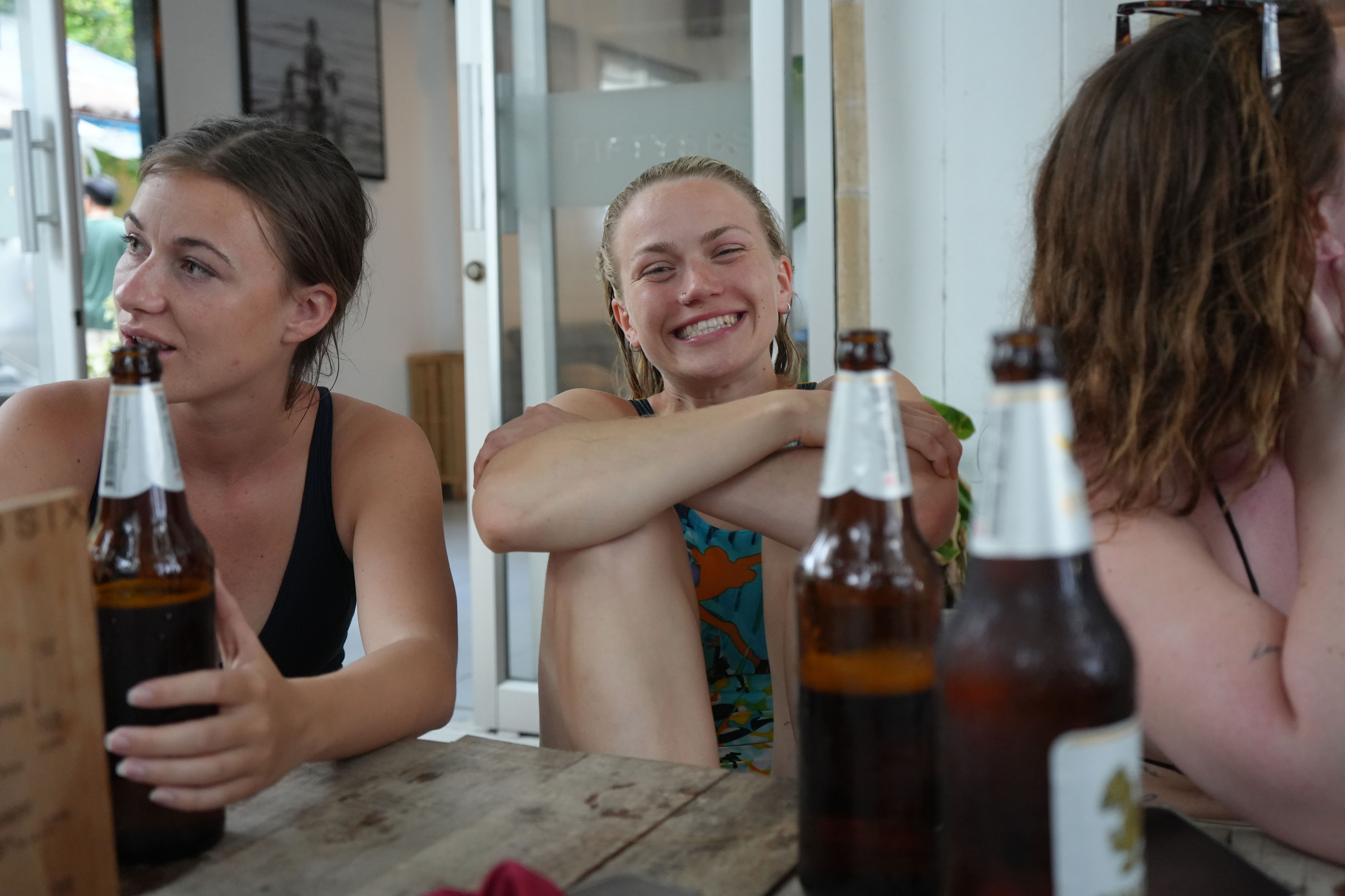A smiling girl sitting at a wooden table with other girls and some beers on it. 
